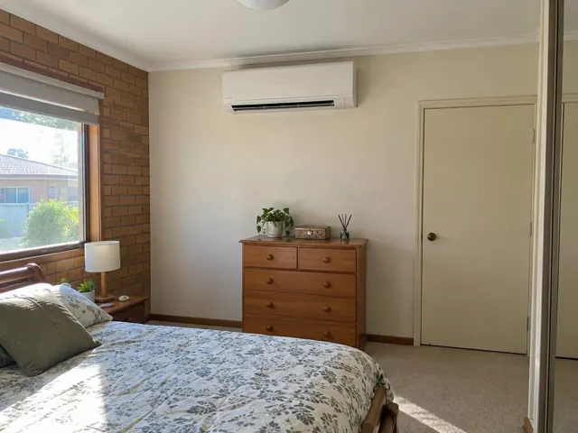 Split system indoor unit mounted high on a bedroom wall above a wooden dresser in a carpeted home in Klemzig